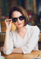 Young woman drinks coffee in cafeteria and posing with sunglasses