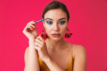 Image of caucasian stylish woman 20s wearing earrings smiling and applying makeup with blue eyeliner, isolated over pink background