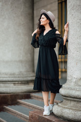 Fototapeta na wymiar Young beautiful stylish girl goes in a black dress in the city. Outdoor summer portrait of a young woman with a hat