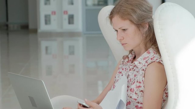 Attractive Businesswoman Sitting On White Chair In Glass Office And Working Behind Laptop