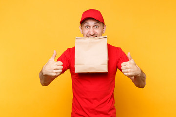 Man in red cap, t-shirt giving fast food order isolated on yellow background. Male employee courier hold in mouth paper packet with food. Products delivery from shop or restaurant to home. Copy space.