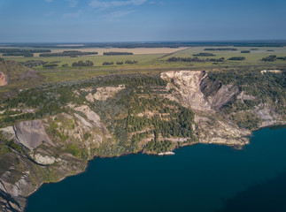 Fototapeta na wymiar Aerial drone view of abandoned quarry with lake appeared on the bottom, the mirror surface of the water in the sunny evening, overgrown with forest shore, meadows on the background, Ural, Russia