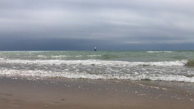 Single windsurfer with black sail far away on waves at sea line, sand under aquamarine water, windy dangerous day, cloudy low sky