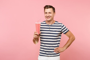 Portrait of young man wearing striped t-shirt holding plastic cup of soda, red glass cola isolated on trending pastel pink background. People youth sincere emotions lifestyle concept. Advertising area