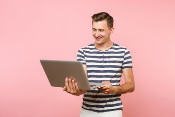 Portrait of handsome young man wearing striped t-shirt working on laptop computer, copy space isolated on trending pastel pink background. People sincere emotions lifestyle concept. Advertising area.