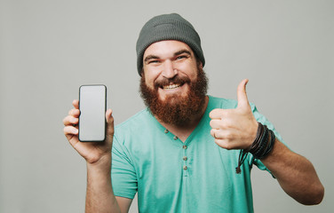 Happy man with beard showing smartphone and thumbs up gesture