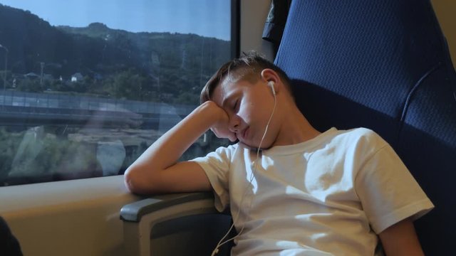 The boy is sleeping by the window in the train car. The boy is traveling the world. Tourist boy on the train