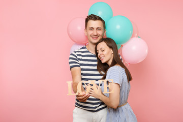 Portrait of young happy smiling couple in love. Woman lean on one's man chest celebrating birthday holiday party on pastel pink background with colorful air balloons. People sincere emotions concept.