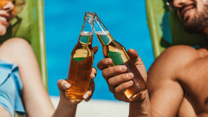 cropped view of couple clinking with bottles of beer on sunbeds