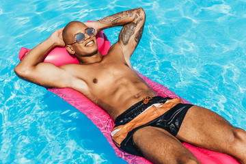 cheerful tattooed african american man sunbathing on pink inflatable mattress in swimming pool