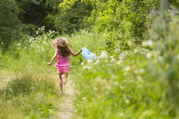 Девочка с длинными светлыми волосами в розовых шортах и футболке с голубой соломенной шляпой в руке бежит в высокой траве