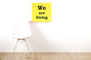Job recruitment advertisement, WE ARE HIRING text written on yellow wall poster, one empty loft style chair. Human resources campaign to find workers for vacant job. Close up, copy space, background.