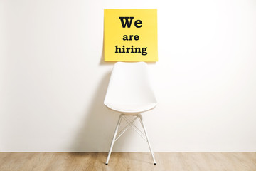 Job recruitment advertisement, WE ARE HIRING text written on yellow wall poster, empty loft style chair. Human resources campaign to find new workers for vacant job. Close up, copy space, background.