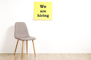 Job recruitment advertisement, WE ARE HIRING text written on yellow wall poster, empty loft style chair. Human resources campaign to find new workers for vacant job. Close up, copy space, background.