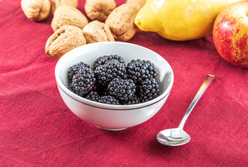 Delicious fresh blackberries and ripe black and reddish garnet. In white bowl on red and white background. With red apple, apricots, lemon and walnuts