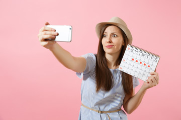 Confused woman in blue dress doing selfie on mobile phone, holding periods calendar for checking menstruation days isolated on pink background. Medical, healthcare, gynecological concept. Copy space.