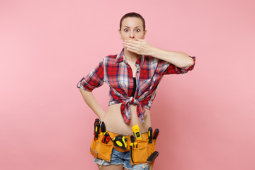 Handyman woman in shirt, denim shorts, kit tools belt full of variety useful instruments cover mouth with hand isolated on pink background. Female doing male work. Renovation and occupation concept.