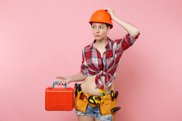 Strong young handyman woman in orange helmet, plaid shirt, denim shorts, kit tools belt full of instruments, toolbox isolated on pink background. Female in male work. Renovation and occupation concept