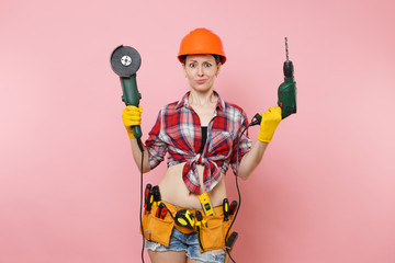 Energy fun handyman woman in gloves, protective orange helmet, kit tools belt full of instruments holding power saw electric drill isolated on pink background. Female in male work. Renovation concept.