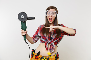 Strong young energy handyman woman in plaid top shirt, denim shorts, kit tools belt full of instruments in protective goggles hold power electric saw isolated on white background. Female in male work.