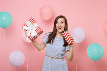 Portrait of joyful beautiful woman in blue dress holding red box with gift present drinking soda or...