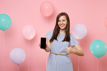 Portrait of beautiful young happy woman wearing blue dress holding mobile phone with blank empty screen showing thumb up on pink background with colorful air balloons. Birthday holiday party concept.