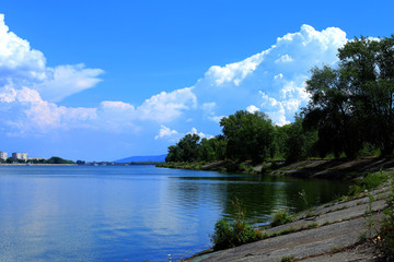 Obraz premium Bright beautiful clouds. The shore of the river, on the horizon is the city and port. A warm summer of nature's beauty.