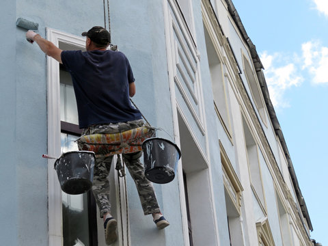 Worker paints the building wall. Painter hanging on a cable with paint buckets, steeplejack repairing house facade with roller brush, renovating works