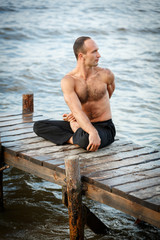 Young yoga trainer practicing twist yoga exercises on a wooden pier on a sea or river shore