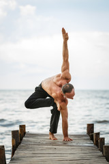 Young yoga trainer practicing yoga exercises on a wooden pier on a sea or river shore.