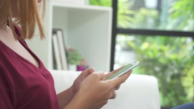 Young asian woman using white smartphone device with green screen. Asian woman mobile , scrolling pages while sitting on the couch in the living room. Chroma key.