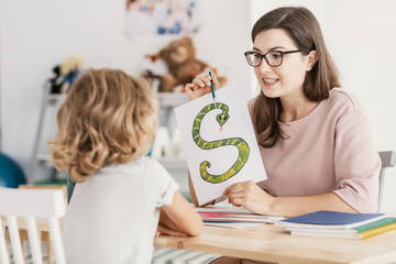A child with development problems with a professional speech therapist during a meeting. Tutor holding a prop poster of a snake as a letter 's'. - obrazy, fototapety, plakaty