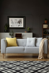 Blanket on grey settee with yellow cushion in living room interior with carpet and poster. Real photo