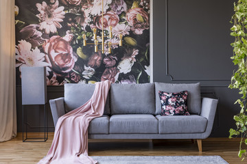 Powder pink blanket thrown on grey couch in real photo of dark sitting room interior with floral wallpaper, gold lamp and wainscoting on wall - obrazy, fototapety, plakaty