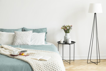 Bed with tea cup, apples and blueberries on breakfast tray and open book in real photo of bright bedroom interior with metal bedside table with flowers