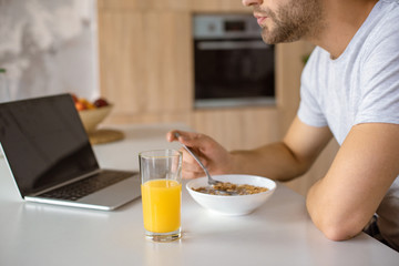 Fototapeta na wymiar cropped image of man eating flakes at kitchen table with fresh juice and laptop