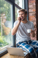 smiling young man with coffee cup talking on smartphone while sitting on windowsill at home