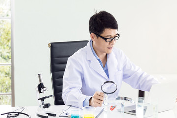 Portrait of a smiling mature scientist in a white coat with magnifying glass near microscope,laptop,test tube at the white lap.Copy space