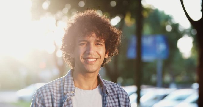 Young south asian guy with curly hair standing on street, smiling and looking at camera - closeup 4k portrait
