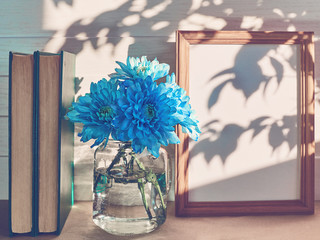 A bouquet of blue chrysanthemums is in a glass jar in the center of the composition. Nearby is a large wooden frame. There are two books. White wooden background with a shadow of a tree on it.
