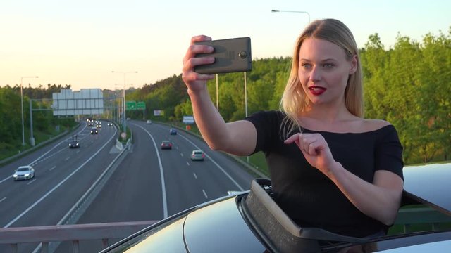 A young beautiful woman stands through a sunroof in a car and takes selfies with a smartphone - a highway in the background