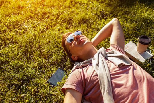 Handsome college man chilling in spring campus park. Happy guy student lying on grass with his phone and coffee beside