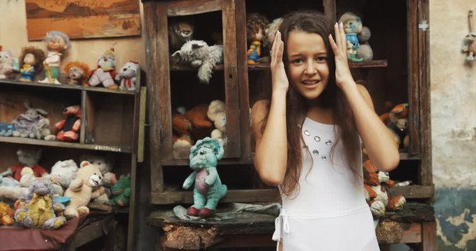 Little girl with dark long hair is hsouting in the garden full of toys and old furniture. 4k footage. Close-up portrait.