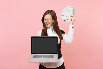 Young laughing business woman holding bundle lots of dollars, cash money and laptop pc computer with blank empty screen isolated on pink background. Lady boss. Achievement career wealth. Copy space.