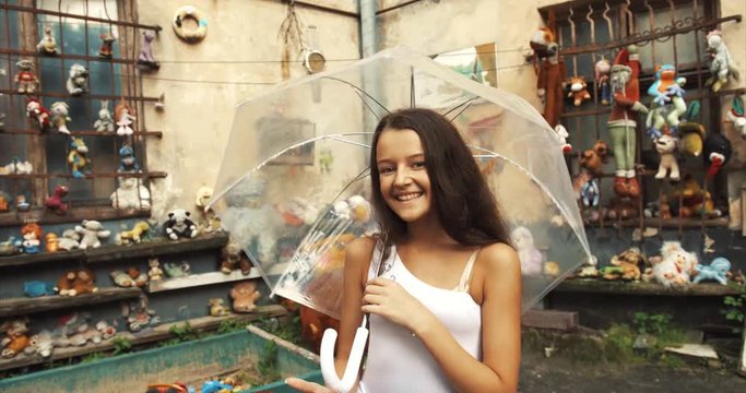 Pretty cheerful little girl with pretty smile and long dark hair having fun in garden full of old toys. She is spinni ng the transparent umbrella. 4k.
