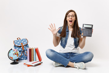 Young shocked excited woman student holding calculator spreading hands learning math sitting near globe, backpack, school books isolated on white background. Education in school university college.