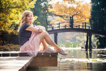 dreaming young woman sit on a wooden pier near lake in autumn city park in a warm fall sunny day