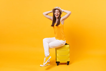 Traveler tourist woman in summer casual clothes, hat sit on suitcase isolated on yellow orange background. Female passenger traveling abroad to travel on weekends getaway. Air flight journey concept.