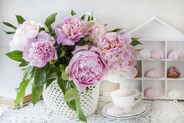 a bouquet of pink peonies on a wooden table in a handmade basket on lace tablecloth, a book, a cup and cakes and meringues