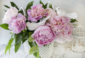 a bouquet of pink peonies on a wooden table in a handmade basket on lace tablecloth, two white birds and a decorative white cage